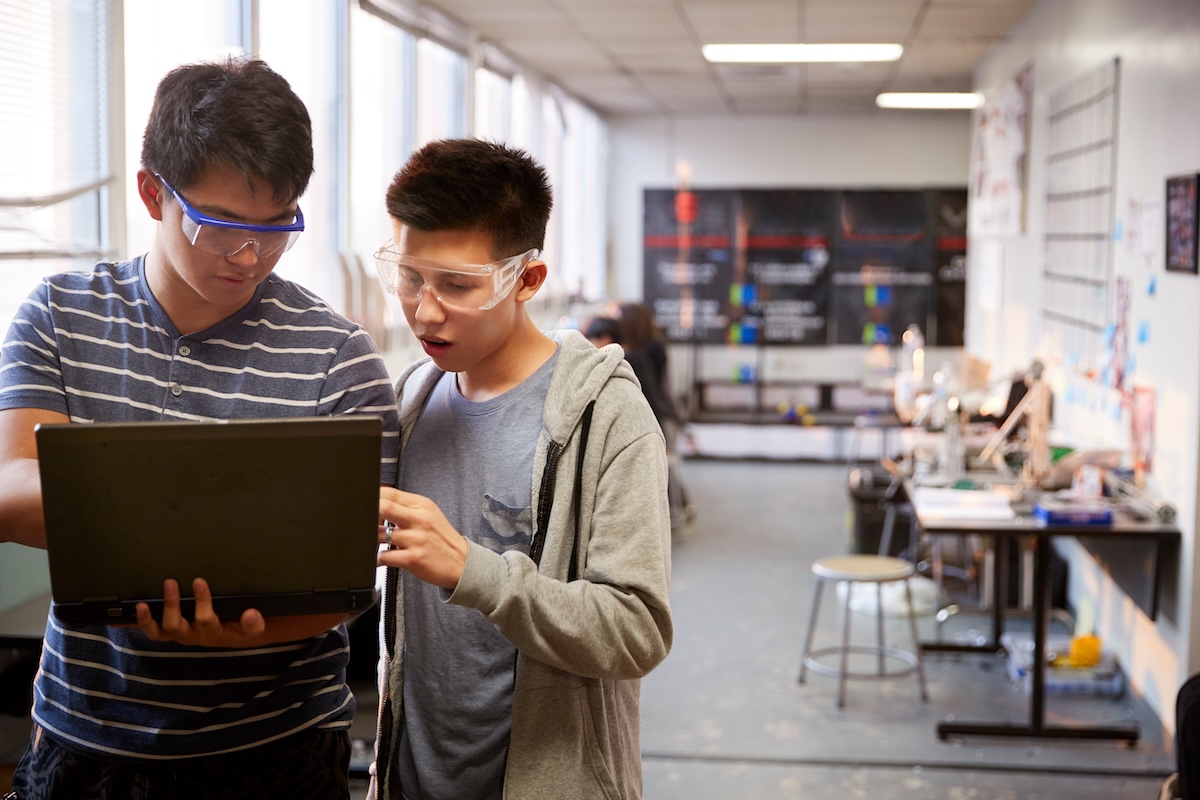 Two students wearing safety goggles using a laptop in a science classroom